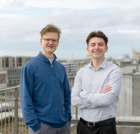 Pascal Nobereit und Dustin Klepper entwickeln bei neuraflow KI-basierte Kommunikationslösungen in Bremerhaven | © Antje Schimanke Zwei junge Männer lächeln draußen an eine Brüstung gelehnt in die Kamera, im Hintergrund ist Wasser und ein verschwommen ein Schiff sowie ein segelartiges Gebäude zu sehen | © Antje Schimanke