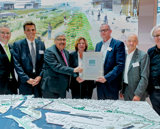 Verleihung des Platin-Vorzertifikats für das LUNEDELTA durch die DGNB im Rahmen der Expo Real | © BIS/Frank Pusch Sechs Männer und eine Frau lächeln in die Kamera, zwei Männer halten ein DIN A4 großen Zettel in die Kamera. Im vordergrund ist ein Modell einer Stadt zu sehen. | © BIS/Frank Pusch